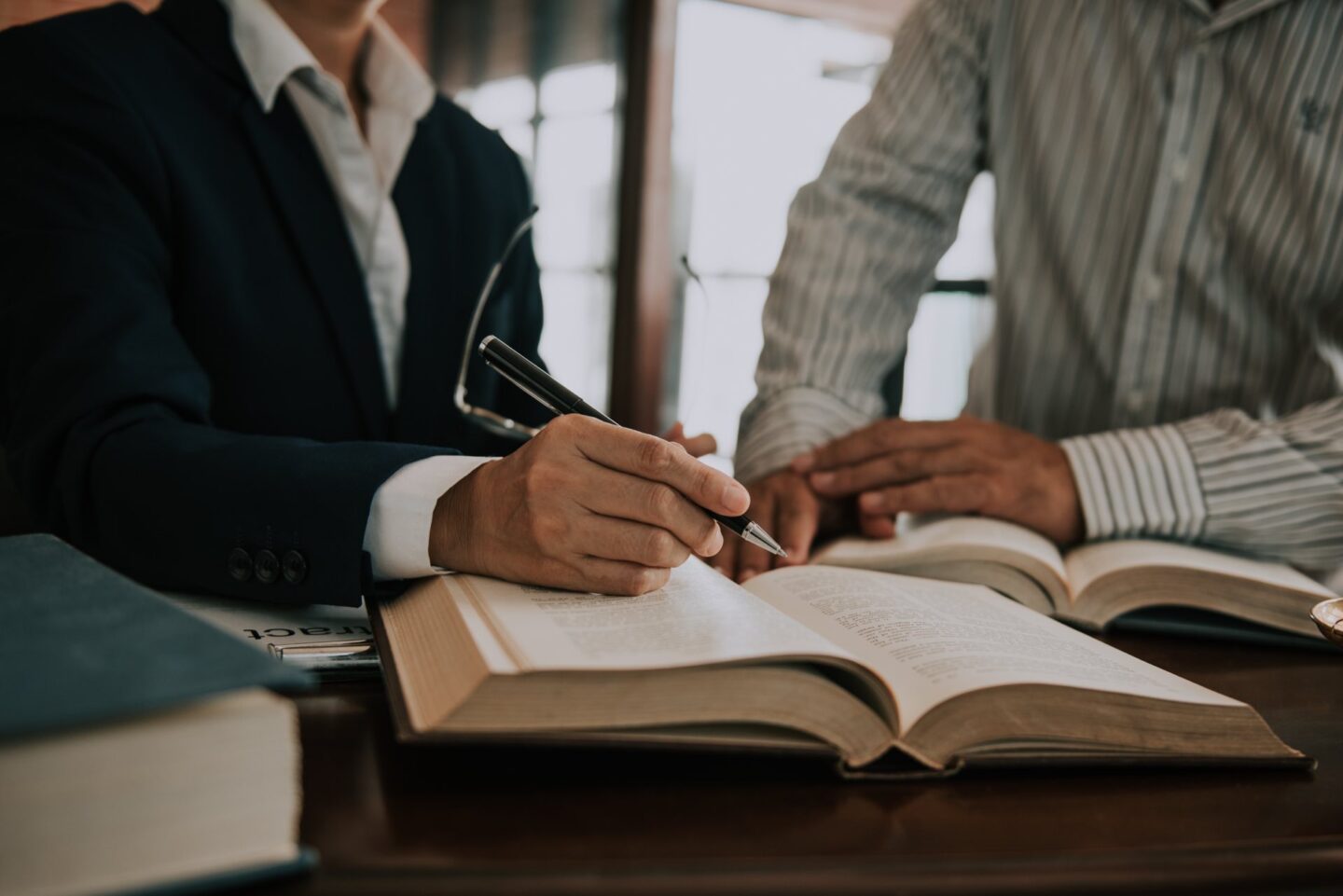 Two people sitting at a desk with books one person holding a pen and pointing at the book